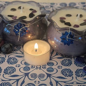 May include: Two decorative candles in blue glass bowls with white wax, adorned with stones and a blue floral embellishment. A lit tea light candle sits in front, with blueberries and buttons scattered around. The candles have a wavy rim.
