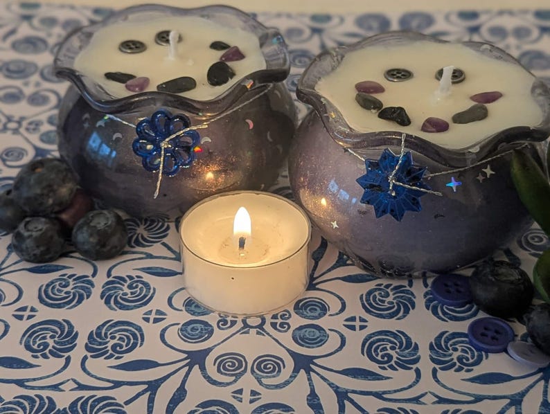 May include: Two decorative candles in blue glass bowls with white wax, adorned with stones and a blue floral embellishment. A lit tea light candle sits in front, with blueberries and buttons scattered around. The candles have a wavy rim.