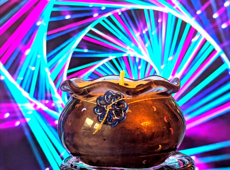 May include: A brown glass candle holder with a yellow candle. The holder has a wavy rim and a blue flower-shaped decoration. The background features a colorful, abstract design with pink, blue, and white lines.
