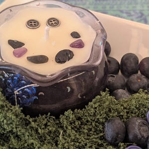 May include: A decorative candle in a clear glass container with a black base, decorated with a floral design and crescent moons. The candle is cream-colored and adorned with small stones and two silver buttons. The candle sits on green moss, surrounded by blueberries and a purple button.