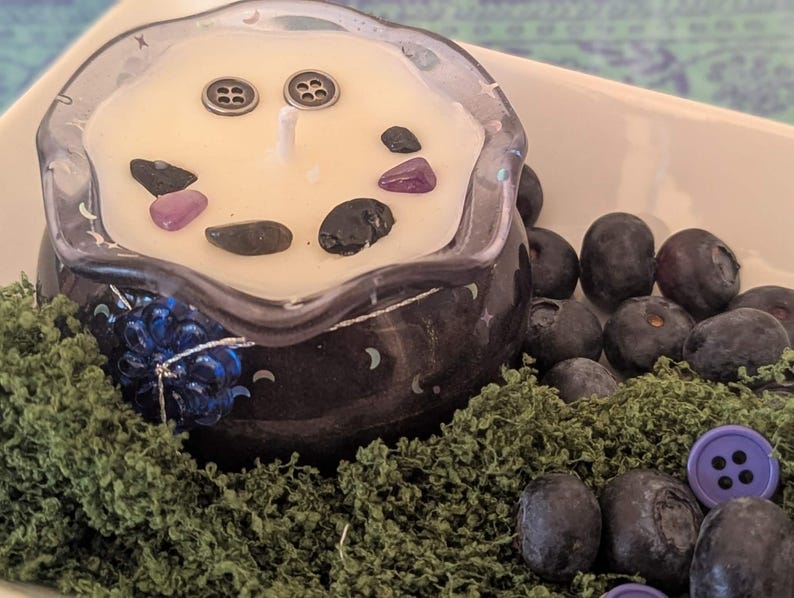 May include: A decorative candle in a clear glass container with a black base, decorated with a floral design and crescent moons. The candle is cream-colored and adorned with small stones and two silver buttons. The candle sits on green moss, surrounded by blueberries and a purple button.