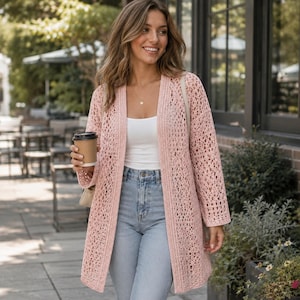Könnte beinhalten: Ein hellrosa, offen gewebter Häkelcardigan. Der langärmlige Pullover ist knielang und wird über einem weißen Tanktop und einer blauen Jeans getragen. Die Person hält eine Kaffeetasse und trägt eine Halskette.