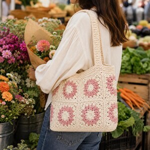Puede incluir: Bolso tote de crochet color crema con dise&ntilde;os florales rosas. El bolso tiene forma rectangular y un asa resistente. El bolso lo lleva una persona en un mercado de flores, rodeada de flores frescas y productos.