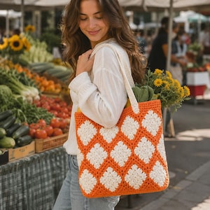 Puede incluir: Un bolso tote de crochet naranja y blanco con un patrón de diamantes se lleva sobre el hombro. La bolsa está llena de comestibles, incluyendo flores amarillas y verduras verdes. La bolsa tiene una correa color crema.
