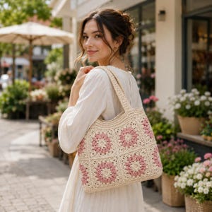 Puede incluir: Un bolso tote de crochet color crema con cuadrados florales rosas. El bolso tiene un asa resistente y se lleva al hombro. El fondo muestra una florister&iacute;a con una mujer con un vestido blanco.