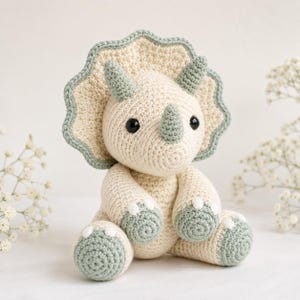 Puede incluir: Un juguete de dinosaurio Triceratops tejido a ganchillo en crema y verde salvia. El peluche hecho a mano tiene un volante, cuernos y pies en verde salvia, con ojos de seguridad negros. El dinosaurio está sentado, mostrando su detallado trabajo de ganchillo.