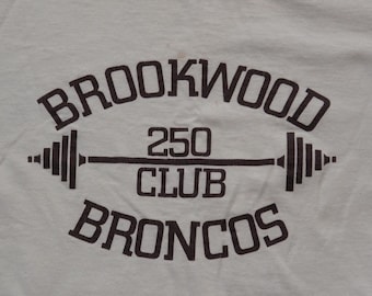 Könnte beinhalten: Weißes T-Shirt mit braunem Aufdruck. Der Aufdruck lautet "BROOKWOOD 250 CLUB BRONCOS" mit einem Hantel-Design. Das Shirt scheint aus Baumwolle oder einem ähnlichen Stoff zu sein.