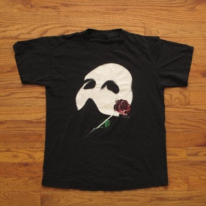 May include: Black t-shirt featuring a white mask and a red rose with a green stem. The graphic design is centered on the front of the shirt. The shirt is laid flat on a wooden surface.