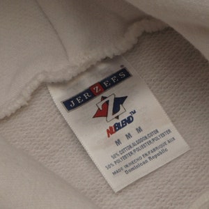 May include: Close-up of a white hoodie with a tag. The tag reads "JERZEES" and "NuBlend" with a red and blue logo. The tag also indicates the hoodie is made of a cotton and polyester blend, size M, and made in the Dominican Republic.