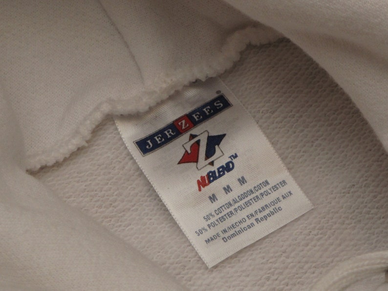 May include: Close-up of a white hoodie with a tag. The tag reads "JERZEES" and "NuBlend" with a red and blue logo. The tag also indicates the hoodie is made of a cotton and polyester blend, size M, and made in the Dominican Republic.