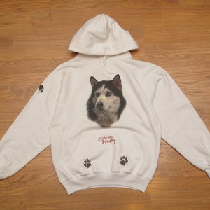 May include: White hoodie featuring a Siberian Husky dog portrait on the front. The design includes the text "Siberian Husky" in red script below the dog's image, and paw prints on the front pocket. A small dog image is on the left sleeve.