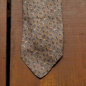 Pode incluir: Uma gravata castanha com estampa paisley com um padrão repetido de pequenos desenhos em forma de lágrima em tons de azul, vermelho e branco. A gravata é feita de um tecido liso e é exibida em uma superfície de madeira.