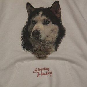 May include: White hooded sweatshirt featuring a detailed graphic of a Siberian Husky's face. The dog has black and white fur with blue eyes. The text "Siberian Husky" is printed in red script below the graphic.