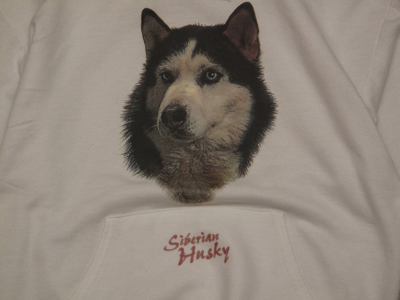 May include: White hooded sweatshirt featuring a detailed graphic of a Siberian Husky's face. The dog has black and white fur with blue eyes. The text "Siberian Husky" is printed in red script below the graphic.