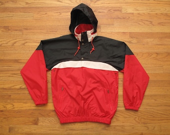 Puede incluir: Una chaqueta anorak con capucha en rojo, negro y blanco. La chaqueta tiene una capucha y una sección superior negras, una franja blanca en el pecho y una sección inferior roja. Cuenta con un cordón, cierres a presión y dos bolsillos delanteros.