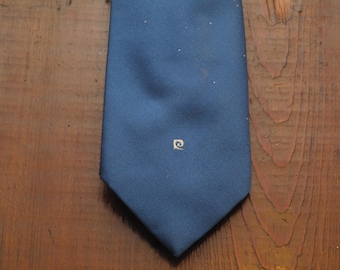 May include: A navy blue tie with a small, square logo near the bottom. The tie is made of a smooth fabric and is displayed on a wooden surface. The logo is a white, stylized design.
