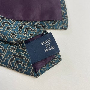 Puede incluir: Primer plano de una corbata estampada en verde azulado y negro con forro morado oscuro. La etiqueta dice "MADE BY HAND". La corbata tiene un dise&ntilde;o geom&eacute;trico y probablemente est&aacute; hecha de seda o un material similar.