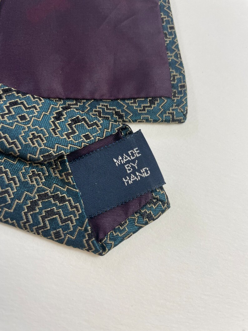 Puede incluir: Primer plano de una corbata estampada en verde azulado y negro con forro morado oscuro. La etiqueta dice "MADE BY HAND". La corbata tiene un dise&ntilde;o geom&eacute;trico y probablemente est&aacute; hecha de seda o un material similar.