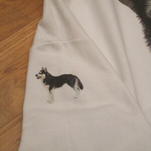 May include: White sweatshirt featuring a detailed illustration of a husky dog on the sleeve. The dog is depicted in black, white, and gray, with a realistic style. The design is on a plain white background.