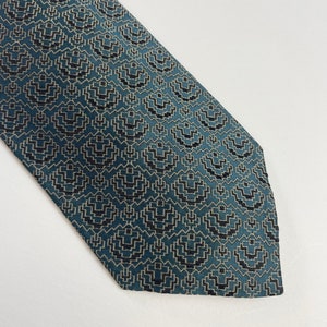 Puede incluir: Una corbata de color turquesa con un patr&oacute;n geom&eacute;trico. El patr&oacute;n consiste en formas negras y blanquecinas repetidas, creando una apariencia texturizada. La corbata est&aacute; en &aacute;ngulo, mostrando su dise&ntilde;o y color.