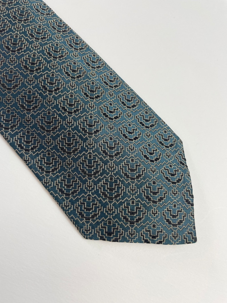 Puede incluir: Una corbata de color turquesa con un patr&oacute;n geom&eacute;trico. El patr&oacute;n consiste en formas negras y blanquecinas repetidas, creando una apariencia texturizada. La corbata est&aacute; en &aacute;ngulo, mostrando su dise&ntilde;o y color.