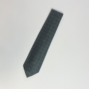 Puede incluir: Una corbata gris con un patr&oacute;n geom&eacute;trico repetitivo en gris oscuro y azul. La corbata est&aacute; en &aacute;ngulo sobre un fondo blanco, mostrando su dise&ntilde;o y textura. Adecuada para vestimenta formal o de negocios.
