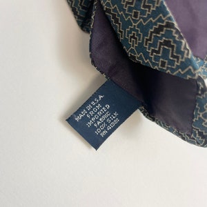 Puede incluir: Primer plano de una corbata de seda estampada. La corbata presenta un dise&ntilde;o geom&eacute;trico en verde azulado, negro y beige. Se ve una etiqueta azul marino, con el texto "MADE IN U.S.A. FROM IMPORTED FABRIC 100% SILK".