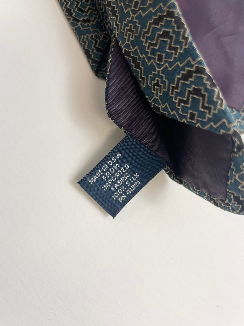 Puede incluir: Primer plano de una corbata de seda estampada. La corbata presenta un dise&ntilde;o geom&eacute;trico en verde azulado, negro y beige. Se ve una etiqueta azul marino, con el texto "MADE IN U.S.A. FROM IMPORTED FABRIC 100% SILK".