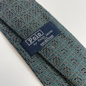 Puede incluir: Primer plano de una corbata estampada. La corbata presenta un dise&ntilde;o geom&eacute;trico en verde azulado, negro y beige. La etiqueta dice "Polo by Ralph Lauren for Nordstrom".