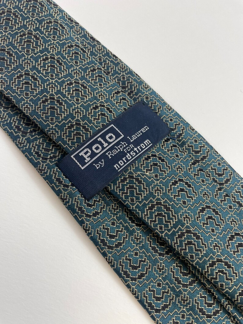Puede incluir: Primer plano de una corbata estampada. La corbata presenta un dise&ntilde;o geom&eacute;trico en verde azulado, negro y beige. La etiqueta dice "Polo by Ralph Lauren for Nordstrom".