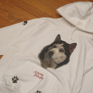 May include: White hooded sweatshirt featuring a detailed portrait of a Siberian Husky with blue eyes. The sweatshirt includes paw prints and the text "Siberian Husky" in red script. The hoodie has a drawstring and a front pocket.