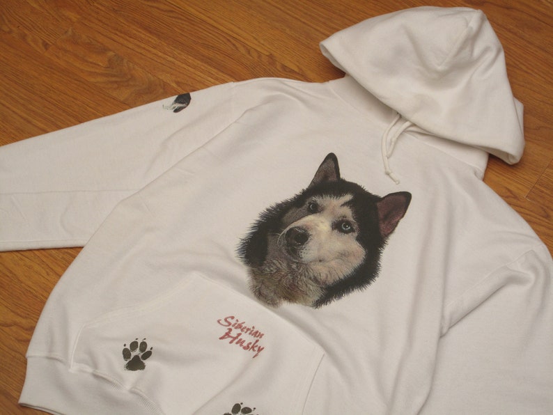 May include: White hooded sweatshirt featuring a detailed portrait of a Siberian Husky with blue eyes. The sweatshirt includes paw prints and the text "Siberian Husky" in red script. The hoodie has a drawstring and a front pocket.