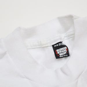 May include: White t-shirt with a Screen Star Best tag sewn into the bottom of the collar. The tag is white with black text and a red star. The tag reads "Screen Star Best" and "Made in USA".