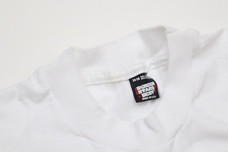 May include: White t-shirt with a Screen Star Best tag sewn into the bottom of the collar. The tag is white with black text and a red star. The tag reads "Screen Star Best" and "Made in USA".