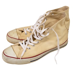 May include: Pair of high-top canvas sneakers in a cream color. The sneakers have white laces, a white rubber toe cap, and a red and blue stripe detail on the sole. The shoes show signs of wear.