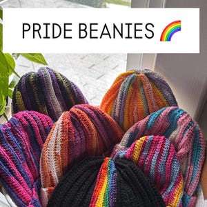 Op de afbeelding: Een verzameling kleurrijke Pride-mutsen in een witte mand. De mutsen zijn in verschillende kleuren, waaronder regenboogstrepen, roze, oranje, paars en blauw. De tekst "PRIDE BEANIES" staat boven de mutsen met een regenboog.