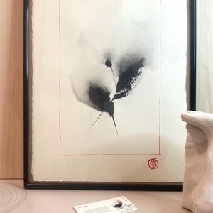 以下が含まれることがあります： 黒いフレームに入った抽象的なインク画。作品は、クリーム色の背景に黒とグレーの花柄のデザインが施され、赤い縁取りがされています。小さなカードと白い彫刻が前景にあります。