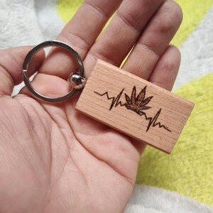 May include: A wooden keychain with a silver key ring. The rectangular wooden piece features a cannabis leaf design and a heartbeat line. The design is etched into the light-colored wood.