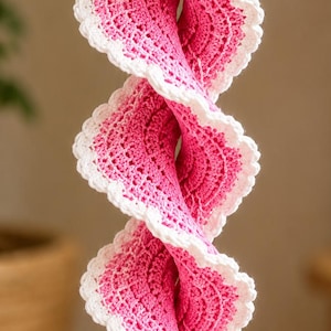 May include: A hanging spiral decoration featuring pink crocheted fabric with a white scalloped edge. The spiral is suspended by a string adorned with pink and clear beads, and it terminates at the bottom with a clear, faceted crystal pendant.