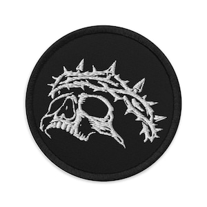 May include: A round, black embroidered patch featuring a white skull wearing a crown of thorns. The skull is detailed with visible eye sockets, teeth, and a defined jawline. The thorns are sharp and pointed, creating a striking contrast against the black background.