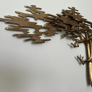 K&ouml;nnte beinhalten: Eine Wandskulptur aus Metall in Messingfarbe, die einen Baum darstellt. Der Baumstamm ist vertikal mit &Auml;sten, die sich nach oben und au&szlig;en erstrecken. Die Bl&auml;tter sind eine Reihe miteinander verbundener, abstrakter Formen. Die Skulptur ist an einer wei&szlig;en Wand befestigt.
