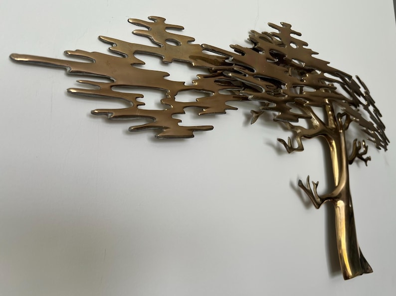 K&ouml;nnte beinhalten: Eine Wandskulptur aus Metall in Messingfarbe, die einen Baum darstellt. Der Baumstamm ist vertikal mit &Auml;sten, die sich nach oben und au&szlig;en erstrecken. Die Bl&auml;tter sind eine Reihe miteinander verbundener, abstrakter Formen. Die Skulptur ist an einer wei&szlig;en Wand befestigt.