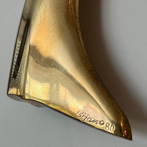 K&ouml;nnte beinhalten: Nahaufnahme einer goldfarbenen, abstrakten Skulptur mit glatter, reflektierender Oberfl&auml;che. Die Skulptur hat eine geschwungene, organische Form. Die Signatur des K&uuml;nstlers, "Bijan 80", ist in die Basis der Skulptur ge&auml;tzt.