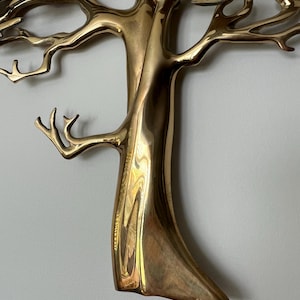 K&ouml;nnte beinhalten: Eine goldfarbene Metallbaumskulptur mit filigranen &Auml;sten und einem strukturierten Stamm. Die Skulptur ist an einer hellen Wand befestigt. Das Design des Baumes ist detailliert, mit poliertem Finish.