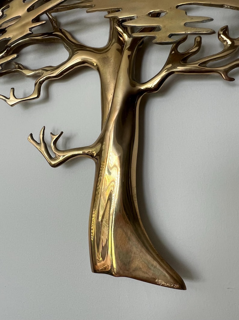 K&ouml;nnte beinhalten: Eine goldfarbene Metallbaumskulptur mit filigranen &Auml;sten und einem strukturierten Stamm. Die Skulptur ist an einer hellen Wand befestigt. Das Design des Baumes ist detailliert, mit poliertem Finish.