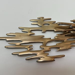 K&ouml;nnte beinhalten: Eine bronzefarbene Metall-Wandskulptur mit einem abstrakten, organischen Design. Das Objekt weist miteinander verbundene, unregelm&auml;&szlig;ige Formen auf, die an flie&szlig;ende Formen erinnern. Die Skulptur ist auf einer wei&szlig;en Oberfl&auml;che montiert.