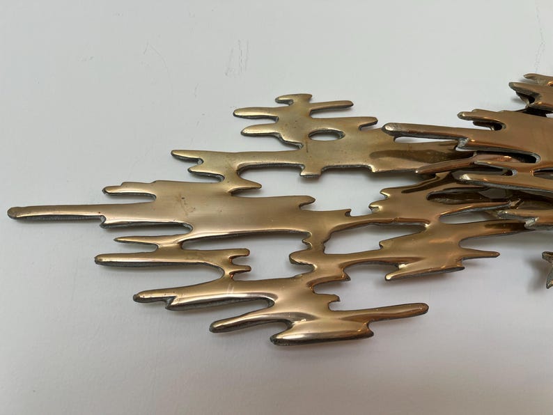 K&ouml;nnte beinhalten: Eine bronzefarbene Metall-Wandskulptur mit einem abstrakten, organischen Design. Das Objekt weist miteinander verbundene, unregelm&auml;&szlig;ige Formen auf, die an flie&szlig;ende Formen erinnern. Die Skulptur ist auf einer wei&szlig;en Oberfl&auml;che montiert.