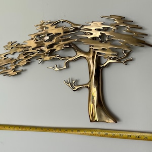 K&ouml;nnte beinhalten: Eine Wandskulptur aus Messing in Form eines Baumes mit detailliertem Design. Der Baum hat einen Stamm und &Auml;ste mit einem strukturierten, geschichteten Bl&auml;tterdach. Ein Ma&szlig;band ist darunter platziert.