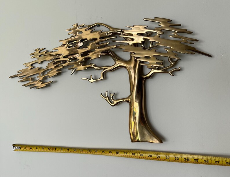 K&ouml;nnte beinhalten: Eine Wandskulptur aus Messing in Form eines Baumes mit detailliertem Design. Der Baum hat einen Stamm und &Auml;ste mit einem strukturierten, geschichteten Bl&auml;tterdach. Ein Ma&szlig;band ist darunter platziert.