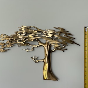 K&ouml;nnte beinhalten: Wandskulptur aus Messing in Form eines Baumes. Der Baum hat einen detaillierten Stamm und &Auml;ste mit einer strukturierten Krone. Die Skulptur ist etwa 35,5 cm hoch. Der Baum ist an einer hellgrauen Wand befestigt.